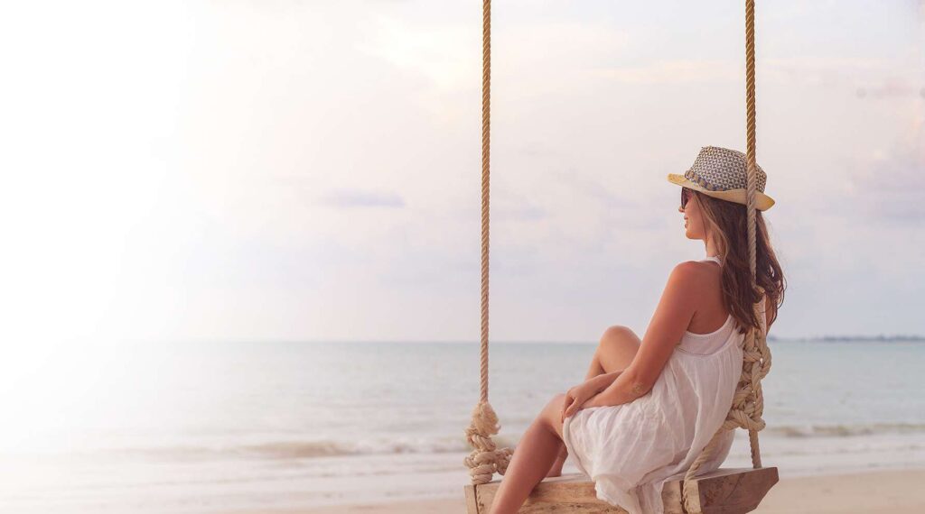 Woman sitting on wooden swing by the sea at sunset