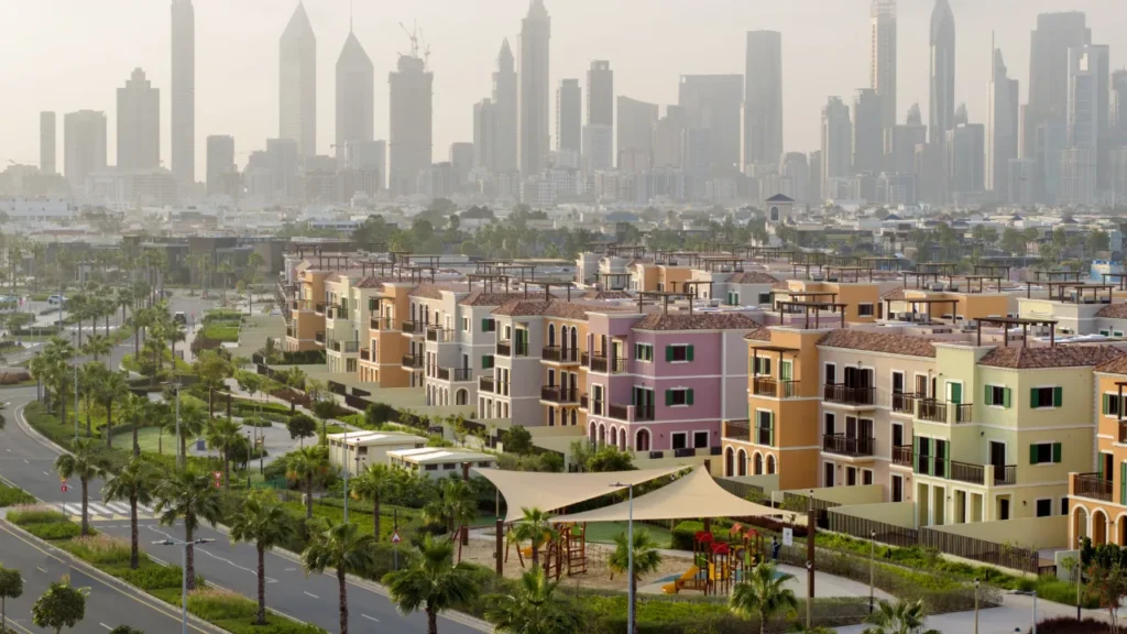 Sur La Mer pastel townhouses with palm-lined street and Dubai high-rises in the haze